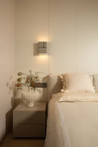 A minimalist bedroom features a side table with a white vase containing a floral arrangement and a bed with light-colored lin