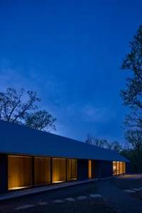 A modern house with wooden exterior and large windows, illuminated from the inside, set against a dark, twilight sky.