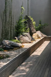 A modern rock garden with large boulders and green plants set against a concrete wall, with a wooden deck in the foreground.