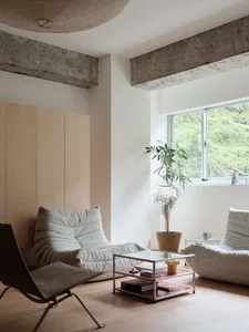 A modern minimalist living room features white sofas, a glass coffee table, and plant accents against a concrete ceiling and 