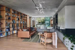 A modern living room with concrete walls, wooden furniture, and a large bookshelf filled with books and various items.