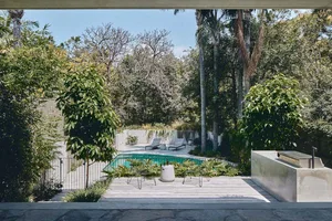 A modern residential backyard featuring concrete patio, pool, and seating area surrounded by greenery and palm trees.