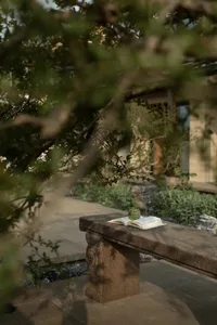 A stone bench with an open book and a fruit resting on top, set in an outdoor space with greenery and a house in the backgrou