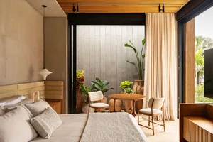 A minimalist bedroom with a bed, chairs, and a small table surrounded by potted plants, featuring hardwood floors and fabric 