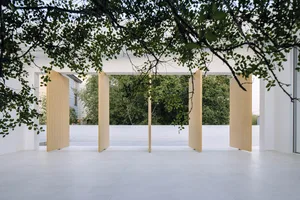 Modern commercial space with large wooden doors and white floors, framed by trees and greenery.