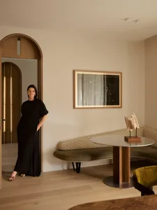 A woman in a black dress standing in a modern living room with fabric upholstery, wooden accents, and metal elements.