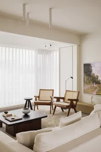 A minimalist living room with two wooden armchairs, a white sofa, a low wooden coffee table, and a large window with sheer cu