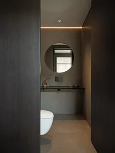 A minimalist bathroom with a white toilet and a circular mirror on a concrete wall.