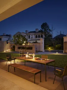 A residential exterior space with wooden and metal elements, featuring a table, chairs, and a bench under warm lighting.