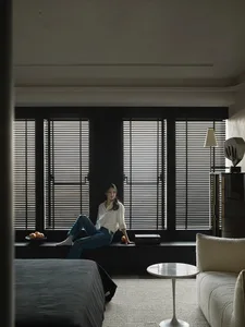 A woman sitting on a window sill in a minimalist bedroom with dark wooden window frames.