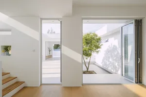 A minimalist white interior space with a large glass window and wooden floor, featuring a tree in the background.