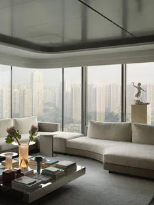 A modern living room with a panoramic city view, upholstered furniture in neutral tones, and a coffee table with books, a vas