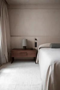 A modern minimalist bedroom featuring a bed with neutral bedding and a wooden nightstand with a lamp.