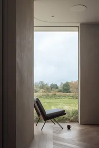 Minimalist interior design featuring a wooden seat, wood flooring, and a large window overlooking a green meadow with trees.