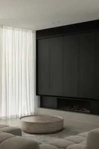 A minimalist living room with white curtains, dark minimalist cabinetry, a circular coffee table, and textured grey seating.