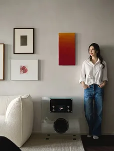 A living room with a modern glass side table, white couch, colorful artwork, and a woman standing against the wall.