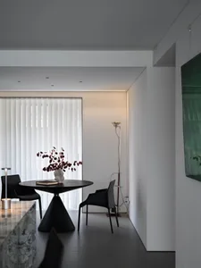 Modern minimalist dining area with a round glass table on a triangular concrete base and black chairs, featuring a vase with