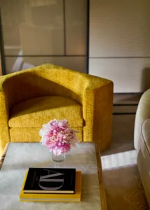 A modern yellow armchair with a marble side table featuring a vase of pink flowers and two books.