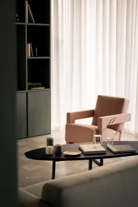 A modern living room with a pink cushioned armchair, a dark coffee table with candles and books, and a bookshelf in the backg
