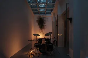 A room with an industrial-style design featuring a drum set, hanging plant, and concrete floor, illuminated by a candle.