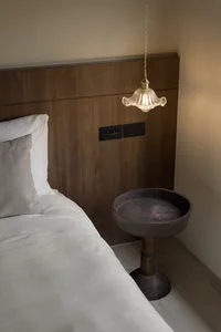 A minimalist bedroom featuring a hanging glass pendant light over a wooden bedside table, with a wooden headboard and white b