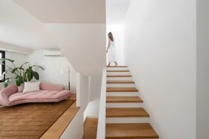 A minimalist interior with a wooden staircase and a blurred person in a white dress walking away.