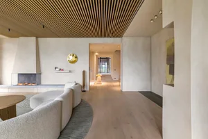 A modern, minimalist living room with white walls, a wood-paneled ceiling, and a fireplace. The space features a comfortable 