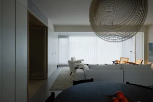 A modern minimalist living room featuring textured white paneling, a black dining table with oranges, and large windows with
