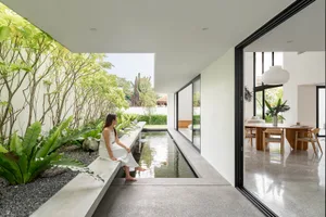 A woman in a white dress sitting in a minimalist indoor garden with a shallow pond and plants