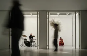 一張極簡室內攝影肖像，描繪音樂家站在門口，前景有模糊的人影走過。