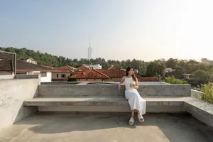 A serene woman sits on a concrete rooftop with a cup in her hand, overlooking a residential neighborhood and a distant skyscr