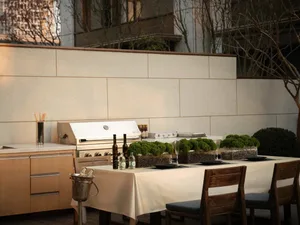 An outdoor kitchen with a concrete countertop and grill, featuring wooden chairs and greenery.