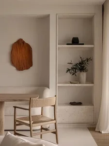 A minimalist living room with a wooden and concrete shelving unit, featuring a potted plant, a vase, and a wooden chair.