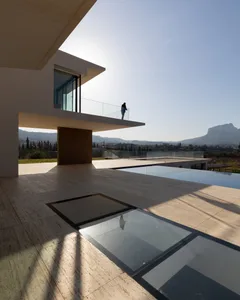 A modern house with glass walls and a concrete patio featuring an infinity pool in an outdoor setting.