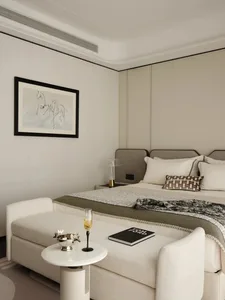 A modern minimalist bedroom with a neutral color palette, featuring a bed with a beige headboard, a matching beige ottoman, a
