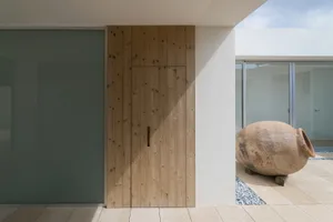 A minimalist wooden door with a handle, set against a concrete exterior wall with a large vase on the floor.
