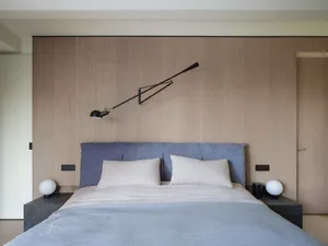 A modern minimalist bedroom featuring a bed with a blue upholstered headboard, white bedding, and a sleek black adjustable re