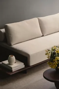 A modern minimalist sofa with beige cushions on a concrete floor with a vase of flowers and a coffee table with books and a v