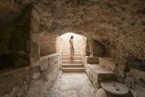 A woman standing in a stone hallway with steps leading up to a brightly lit area.