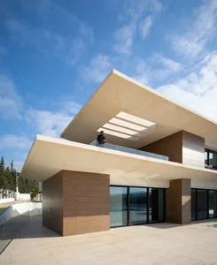 Contemporary house with concrete walls and roof, open space, large windows, and a man standing on the balcony under the roof.