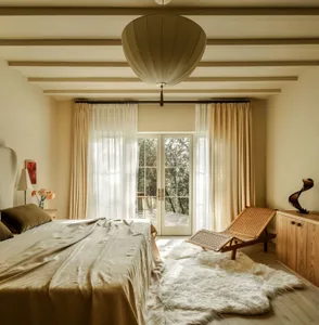 A minimalist bedroom with a bed covered in silk, large windows, and a rattan lounge chair.