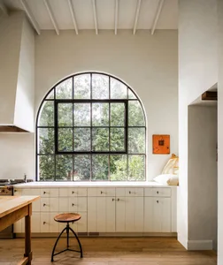 A minimalist kitchen with white cabinets and a large arched window overlooking greenery.