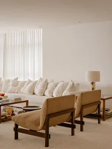 A minimalist living room with neutral tones, featuring a white sofa with multiple cushions, two brown armchairs, and a table