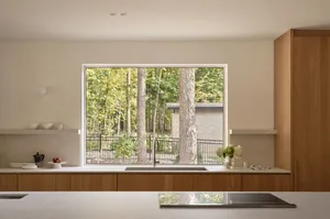 A modern kitchen with wooden cabinets, a sink, and a view through a large window showing a forested area with tall trees.