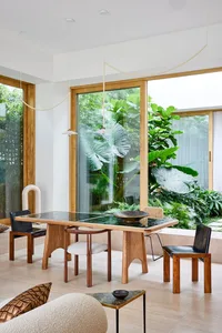 A modern dining room featuring a wooden table with green tiles, surrounded by natural light and lush greenery outside the win