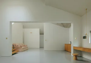 A minimalist living room with concrete and white walls, a low wooden bench, and a beige sofa with pillows.