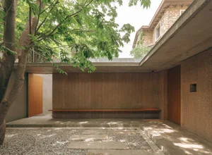 A minimalist residential patio featuring concrete flooring and wooden bench under a tree, with a roof providing shade.