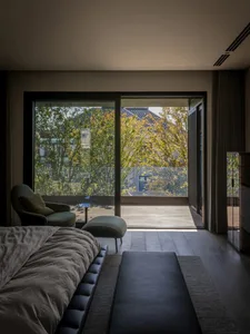 A modern bedroom featuring a large bed, a bench, and a large glass window overlooking trees and a deck.