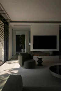 Modern living room with concrete, glass, and upholstered furniture, featuring a large television and a fireplace.