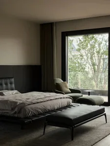 Modern minimalist bedroom featuring a black bed with a leather headboard and footrest, a green armchair, and large windows wi